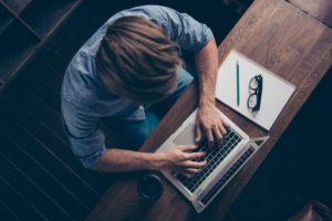 Man sitting at table on a laptop with a notepad and glasses next to him