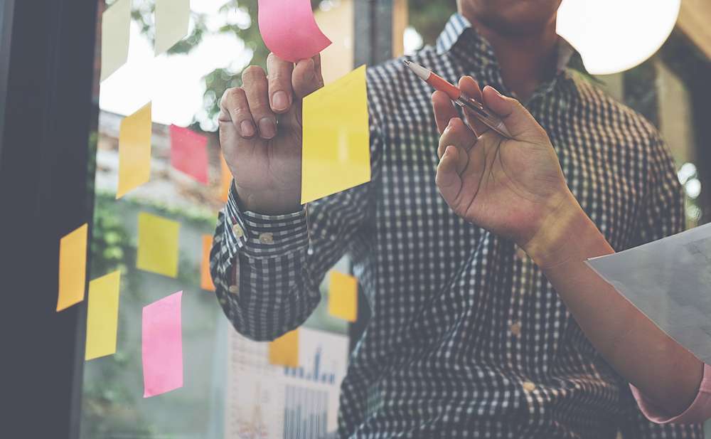 Two co-workers brainstorming, writing on sticky notes, sticking on glass