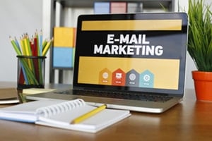 Desk with a notebook, pencils, plant, and a laptop displaying "E-mail Marketing" on it