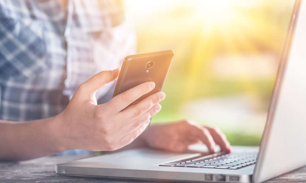 person sitting in front of large laptop, looking at cellphone on a sunny day