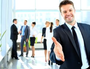 Man wearing a suit sticking hand out for a handshake with business people in background