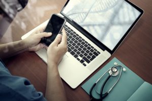 Doctor sitting in front of a laptop, next to a stethoscope, holding a smart phone